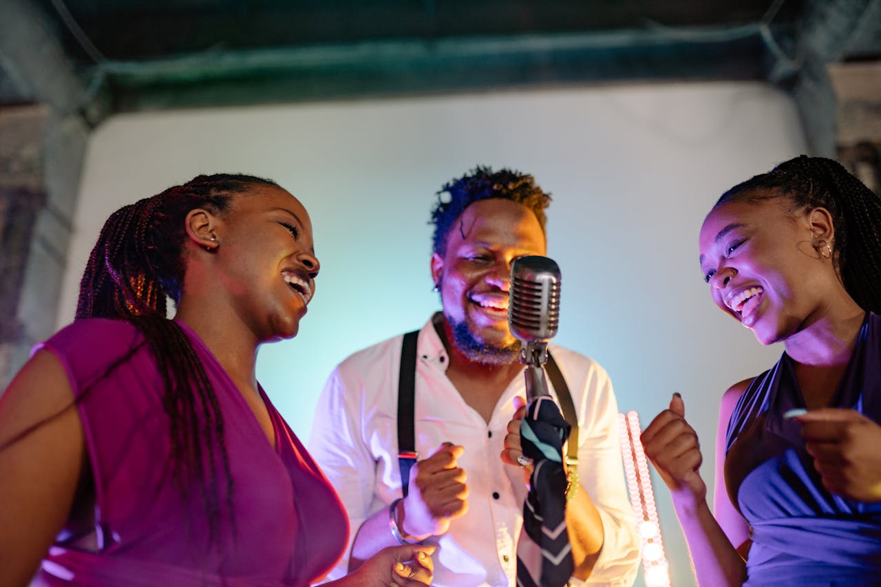 Three smiling adults singing into a microphone under colorful stage lights, exuding joy and energy.